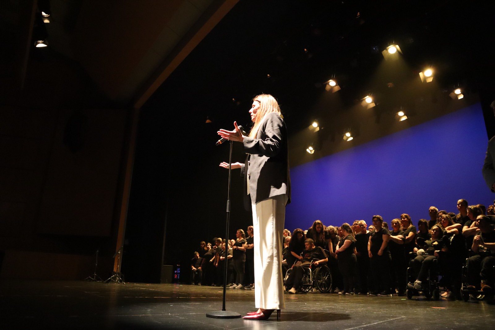 Palco de teatro com iluminação superior direcionada. Em primeiro plano, uma pessoa está de pé junto a um microfone, com um braço levantado e o outro parcialmente aberto, voltada para o público. No fundo do palco, um grupo numeroso de pessoas está alinhado, algumas de pé e pelo menos uma em cadeira de rodas. O cenário é marcado por um fundo liso de cor azul e pela presença de focos de luz no teto.