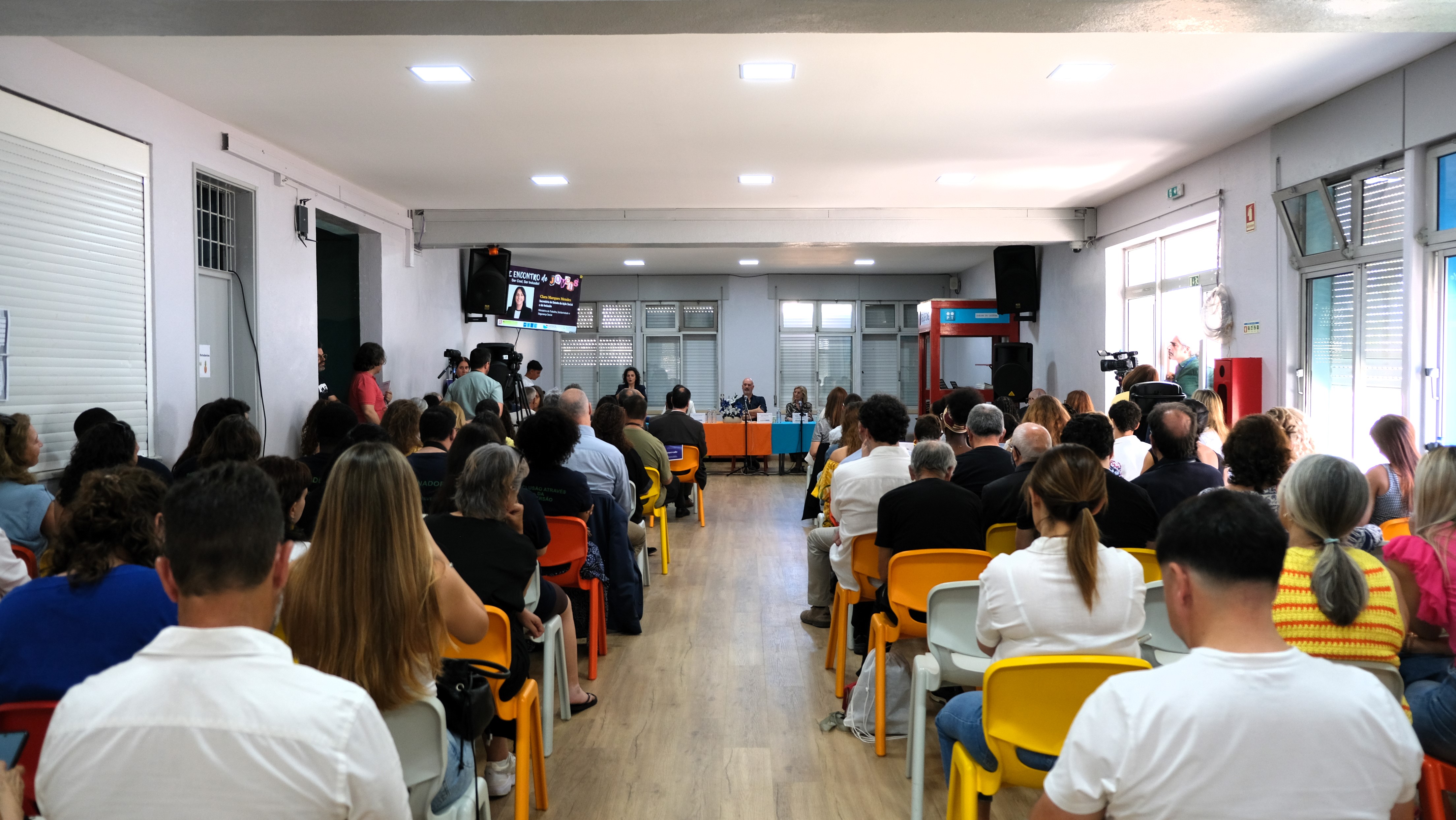  Vista da sala com várias filas de cadeiras ocupadas por participantes voltados para uma mesa onde decorre um painel. Um ecrã está visível na parede ao fundo. View of the room with several rows of chairs occupied by participants facing a table where a panel takes place. A screen is visible on the wall in the background. 