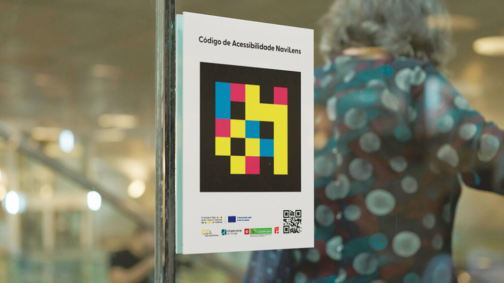  Cartaz colado numa superfície de vidro, visto em contexto interior com fundo desfocado. O cartaz apresenta, na parte superior, o texto “Código de Acessibilidade NaviLens”. Ao centro, encontra-se um código visual composto por vários quadrados de cores contrastantes (amarelo, azul, vermelho e preto), organizados em padrão geométrico sobre fundo preto. Na parte inferior do cartaz, estão vários logótipos de entidades parceiras e um código QR no canto inferior direito. Poster glued to a glass surface, seen in an interior context with blurred background. The poster shows the text “NaviLens Accessibility Code” at the top. In the center, there is a visual code composed of several squares of contrasting colors (yellow, blue, red and black), organized in geometric pattern on a black background. At the bottom of the poster are several partner entity logos and a QR code at the bottom right. 
