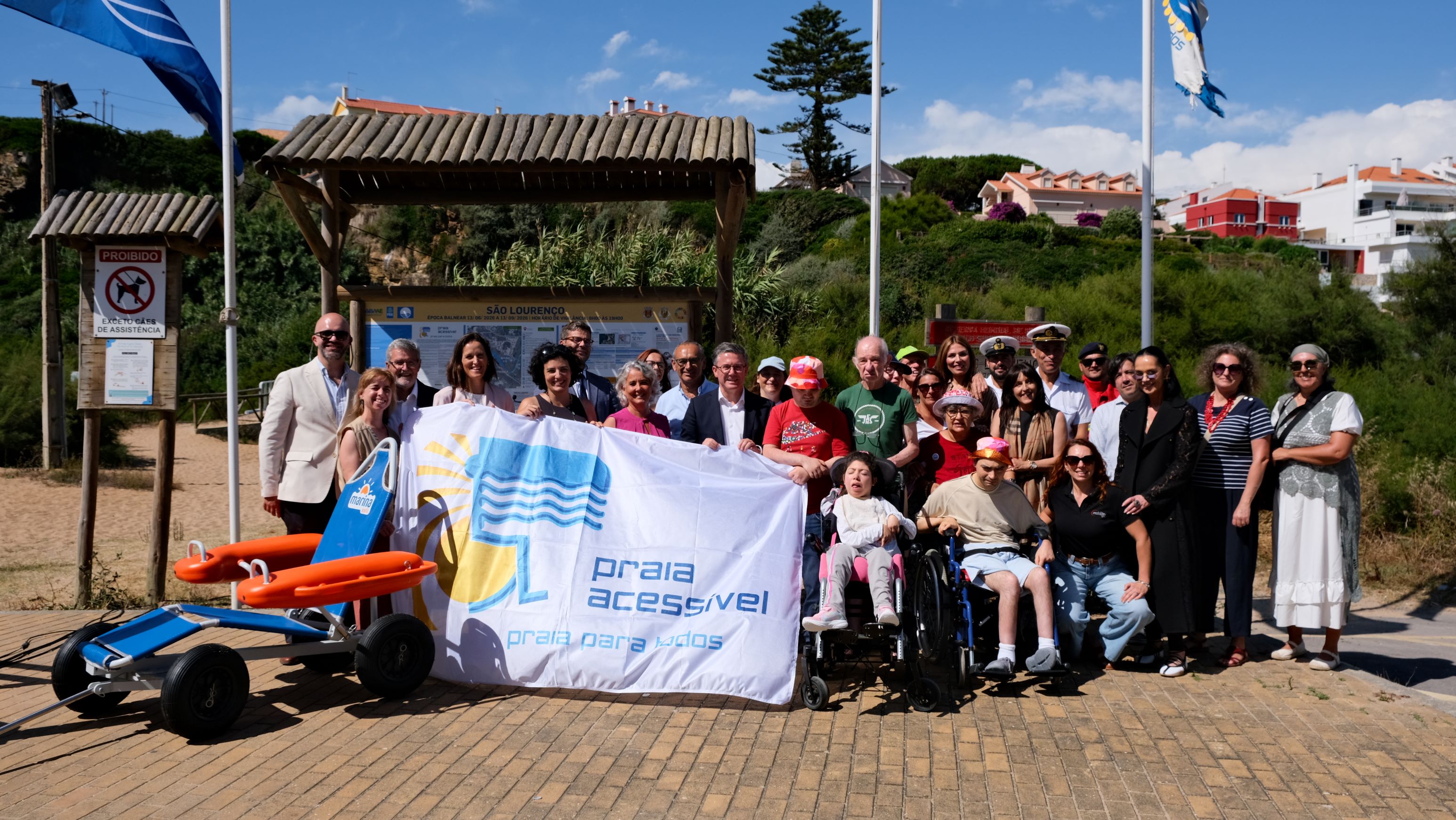  Grupo de cerca de 20 pessoas reunido ao ar livre, em frente a um acesso de praia, a pousar para uma foto. Algumas pessoas utilizam cadeiras de rodas. Parte delas seguram uma bandeira com o fundo branco, com um sol amarelo com raios à esquerda, um ícone azul que combina um símbolo de cadeira de rodas com ondas, representando água ou mar. É possível também ler “praia acessível – praia para todos”. À esquerda, encontra-se uma cadeira anfíbia de praia. Atrás do grupo existem dois mastros com bandeiras e um painel informativo com o título “São Lourenço”. Group of about 20 people gathered outdoors, in front of a beach access, landing for a photo. Some people use wheelchairs. Some of them hold a flag with a white background, with a yellow sun with rays on the left, a blue icon that combines a wheelchair symbol with waves, representing water or sea. You can also read “accessible beach – beach for all”. On the left is an amphibious beach chair. Behind the group there are two flagged masts and an information panel entitled ‘São Lourenço’. 
