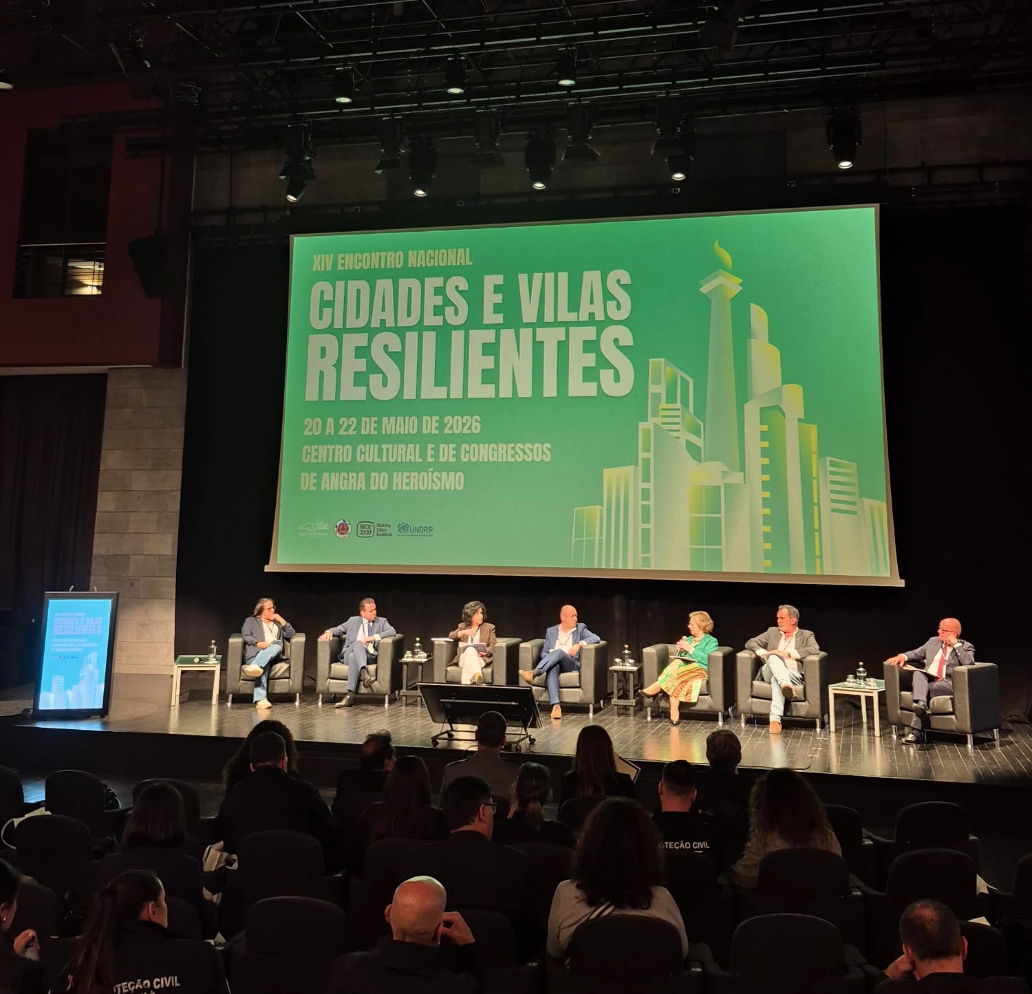  Fotografia de um auditório durante um evento. No palco, várias pessoas estão sentadas em cadeiras dispostas em linha, a participar num painel de debate. À frente do palco encontram-se cadeiras ocupadas pelo público. Ao fundo, um grande ecrã projeta um fundo verde com texto em branco que diz: “XIV Encontro Nacional – Cidades e Vilas Resilientes”. O ecrã inclui ainda a informação “20 a 22 de maio de 2026” e “Centro Cultural e de Congressos de Angra do Heroísmo”. À direita do texto, há uma ilustração estilizada de edifícios urbanos. Photo of an auditorium during an event. On stage, several people are sitting in chairs arranged online, participating in a panel discussion. In front of the stage are chairs occupied by the public. In the background, a large screen projects a green background with blank text that says: “XIV Encontro Nacional – Cidades e Vilas Resilientes”. The screen also includes the information ‘20 to 22 May 2026’ and ‘Centro Cultural e de Congressos de Angra do Heroísmo’. To the right of the text is a stylized illustration of urban buildings. 