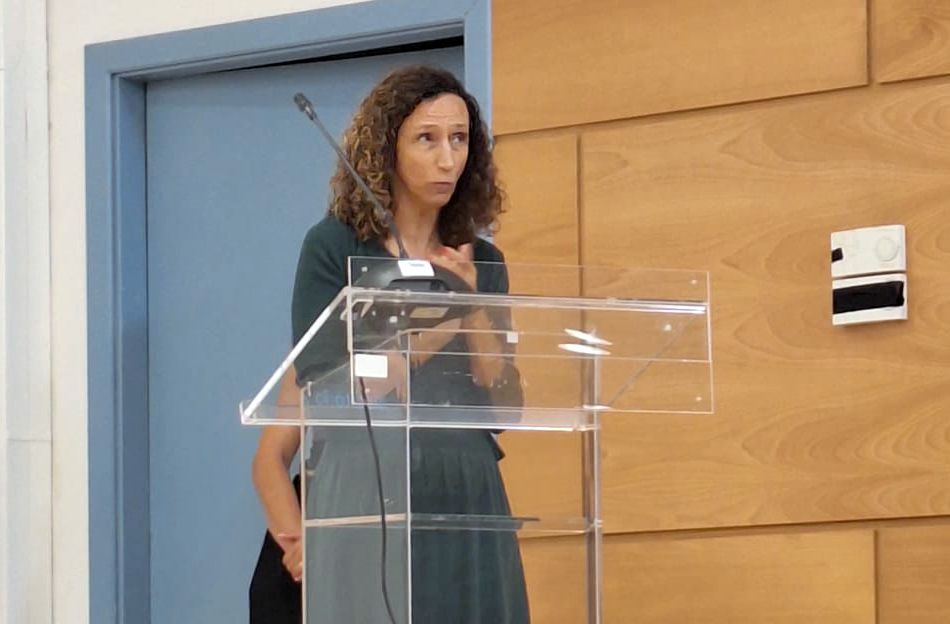 Odete Fiuza encontra-se a discursar atrás de um púlpito transparente numa sala de conferências, durante a sessão de abertura do Mestrado em Atividade Física da Universidade de Leiria e Oeste, em representação do IDiPD. Ao fundo, observam-se painéis de madeira e uma port