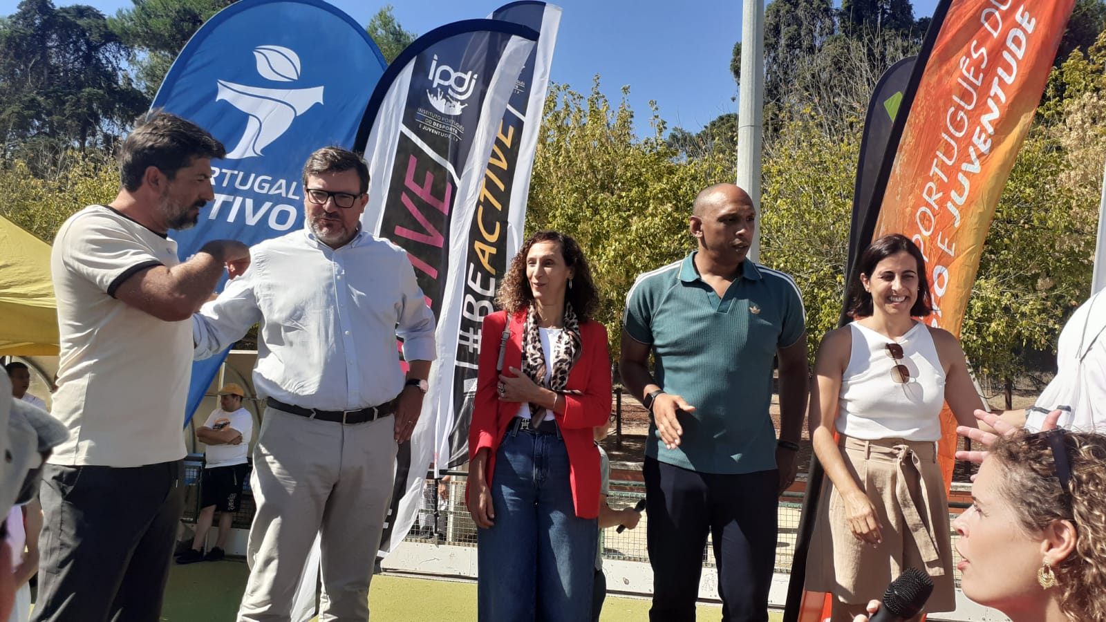  Grupo de participantes ao ar livre durante uma atividade do Dia do Desporto Inclusivo, integrado na Semana Europeia do Desporto. Em destaque, várias pessoas junto a bandeiras institucionais do IPDJ e de iniciativas de promoção da atividade física e juventude. Group of outdoor participants during an Inclusive Sport Day activity as part of the European Week of Sport. Highlighted, several people next to institutional flags of the IPDJ and initiatives to promote physical activity and youth. 
